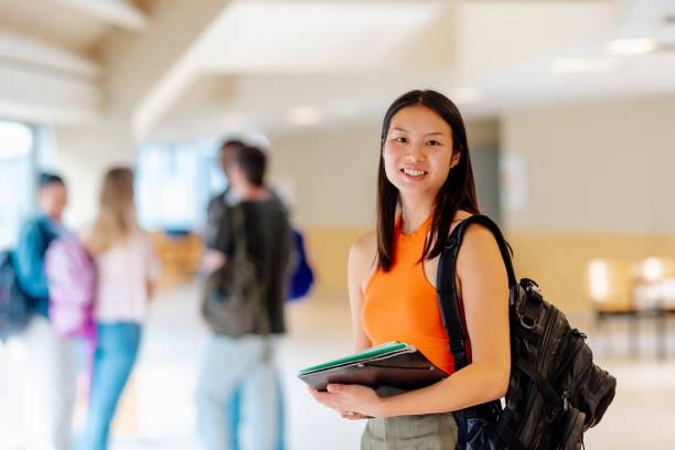 Portrait of an Asian girl with a backpack and school supplies in the hallways of the university campus with her classmates in the background.
